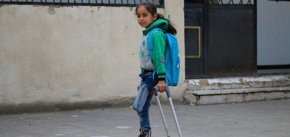 A young girl with one leg is standing in front of a run-down building. She is using a crutch and waering a blue backpack.