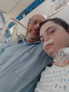 A selfie of a young boy with his father. They are laying in a hospital bed together and the son has a hospital gown on and medical equipment on his face and neck.