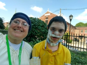A selfie image of a mom and her teenage son. She is wearing a white t shirt, green lanyard, glasses, and a bandana. He is wearing a yellow shirt and has medical tape and equipment on his face and neck.