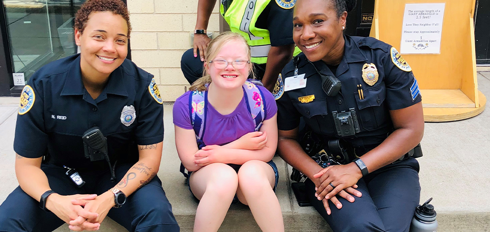 A photo of two woman in their police officer uniforms sitting on either side of a young girl in front of a police station.
