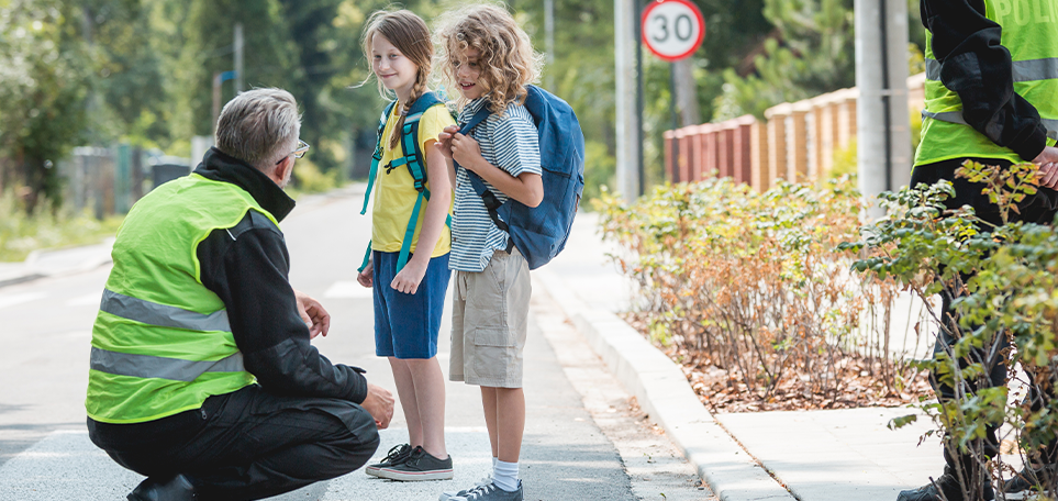 A crossing guard wearing a bright green, reflective vest, kneels down at eye level with two children. Both children are wearing shorts, t-shirts, and have backpacks on. There is a road and sidewalk in the background. Another crossing guard looks on in the background.