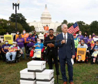 A senator stands in a suit, speaking in front of a group of activists. The US Capitol is behind them, and beside the Senator are 5 large white boxes stacked.