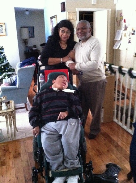 A mom and dad young man who is in wheelchair. They are standing in their living room.