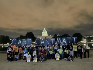 A group of activitists poses in front of the US Capitol at night, holding light up signs that say Care Can't Wait