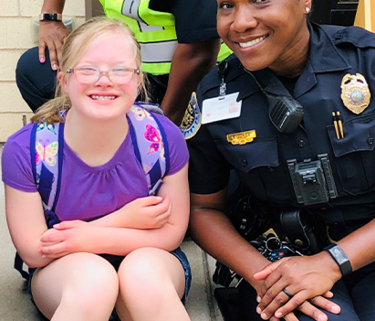 A photo of two woman in their police officer uniforms sitting on either side of a young girl with Down syndrome in front of a police station.