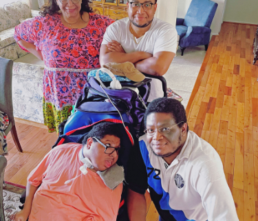 A mom, dad and young man stand around another young man who is in wheelchair. They are standing in their living room.