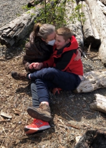 A woman sits on the ground with mulch and a fallen tree around her. On her lap is her young son. She is wearing a mask and holding him affectionately. 