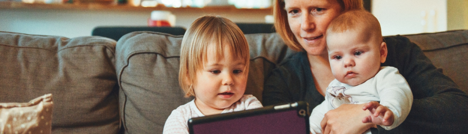 A mother sits on a dark gray couch, holding a baby. A toddler sits next to her holding a tablet, which they are looking at together.