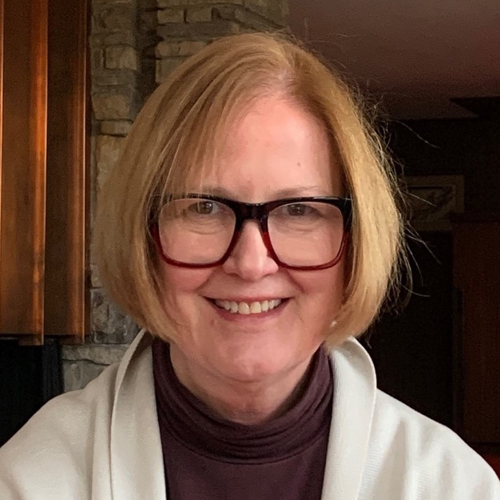A woman with short blonde hair wearing a cream jacket with a burgundy turtleneck underneath and burgundy glasses
