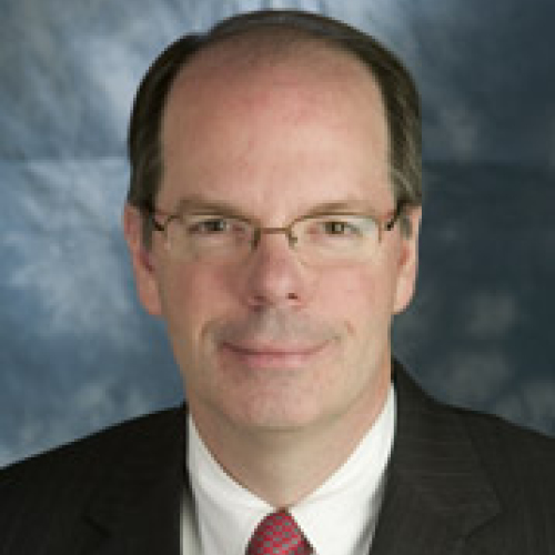 A man with short brown hair wearing a black suit jacket with a white dress shirt and tie and glasses.