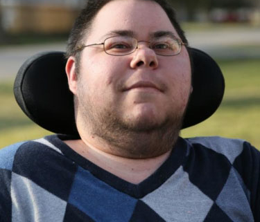 Dominick a masculine appearing person with green eyes and brown spiked short hair gives a small smile. He is wearing gold wire-rim glasses and has some stubble and hair along his cheek line. This headshot shows from his mid-chest area upward. He is wearing a blue, black, and gray Argyle long-sleeve shirt. He sits in his wheelchair, and his black headrest wraps around the back of his head. The background is blurred, but you can tell there is grass behind him and he is outside.