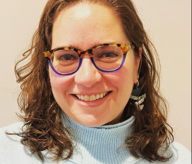 A woman with shoulder length curly brown hair wearing a blue turtleneck sweater and tortoise frame glasses.