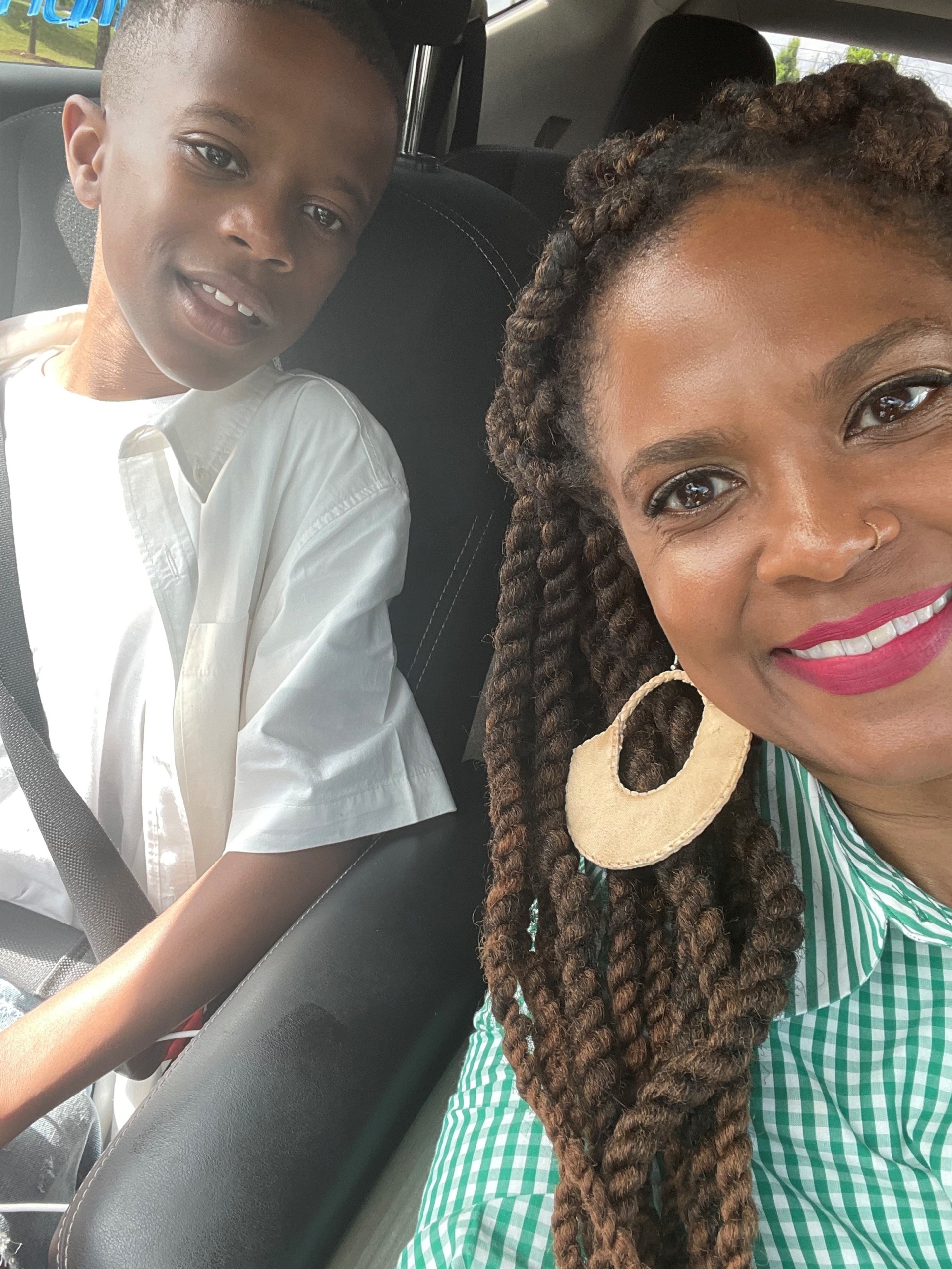 A mom and her son sit in a car, smiling for a selfie. The son is young, and both are Black. The mom is wearing large round white earrings, red lipstick, a green dress, and the son is wearing a white t shirt. 
