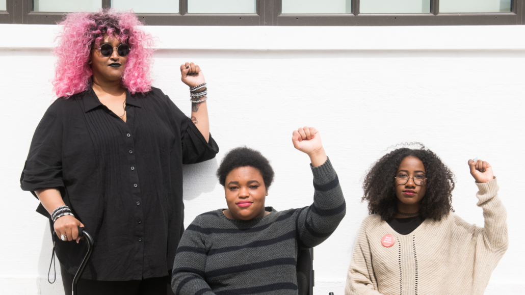Three Black women look resolutely at the camera with their fists raised. Two are seated and one is standing and has pink hair.