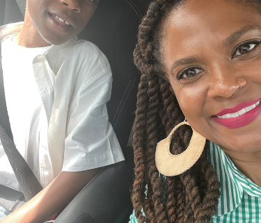 A mom and her son sit in a car, smiling for a selfie. The son is young, and both are Black. The mom is wearing large round white earrings, red lipstick, a green dress, and the son is wearing a white t shirt.