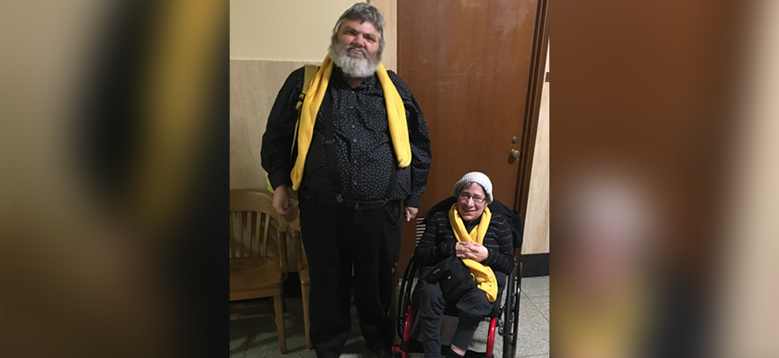 A man with a white beard and wearing all black stands next to a woman in a wheelchair wearing a white stocking hat and wearing all black clothing. They are both have yellow scarves around their necks.