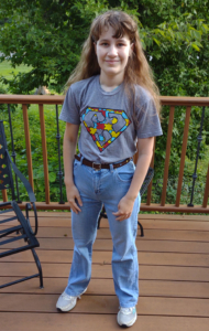 A woman with long brown hair stands on a deck overlooking grass down below. She is wearing a grey t-shirt and jeans.