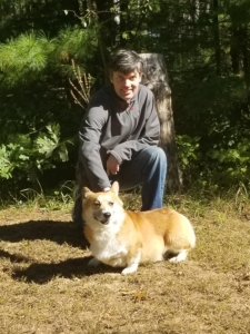 A young man squats, posing with his dog and putting his hand on its head. He is smiling and there are evergreen trees behind them.