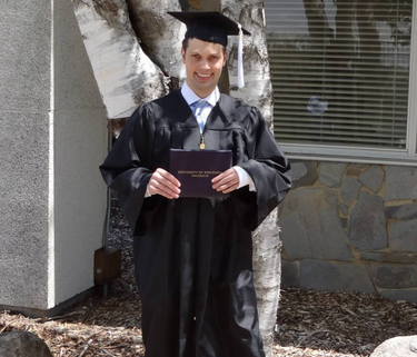A graduate stands in front of a tree on a sandy spot with boulders around him. He is wearing a cap and gown, holding a diploma, and smiling.