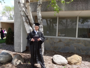 A graduate stands in front of a tree on a sandy spot with boulders around him. He is wearing a cap and gown, holding a diploma, and smiling. 
