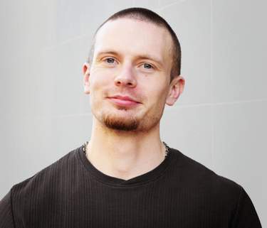 A man smiles softly, wearing a black shirt. His head is shaved and he has a short goatee.