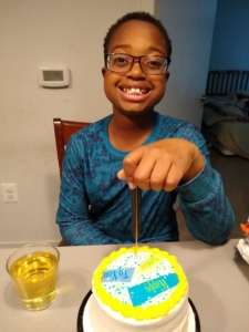 A young boy of color smiles, seated at a table with a birthday cake in front of him. 