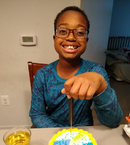 A young boy of color smiles, seated at a table with a birthday cake in front of him.