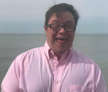 A man in a pink button down stands in front of the ocean, smiling with his eyes closed. He is wearing glasses.