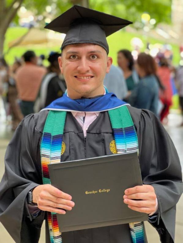 A Hispanic man stands in a black graduation cap and gown, smiling and holding a diploma. He is wearing a multi-colored stole over his gown. 
