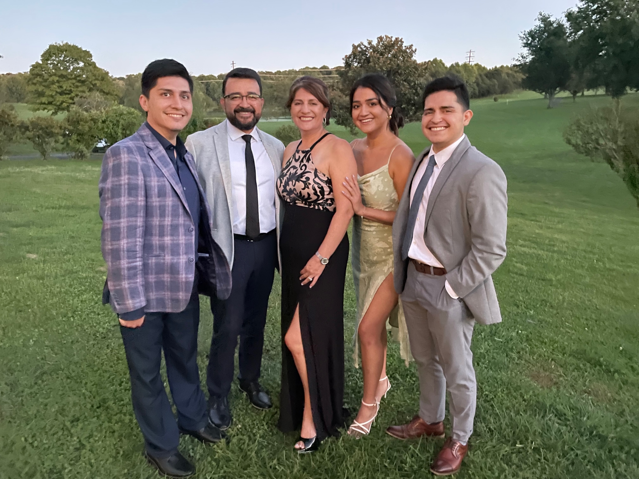 A family including a mom, dad, and three siblings stands posing together and smiling in formal wear. They are standing on a grassy field with trees in the background.