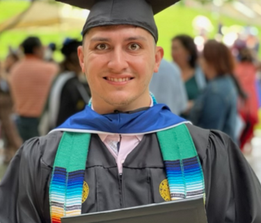 A Hispanic man stands in a black graduation cap and gown, smiling and holding a diploma. He is wearing a multi-colored stole over his gown.