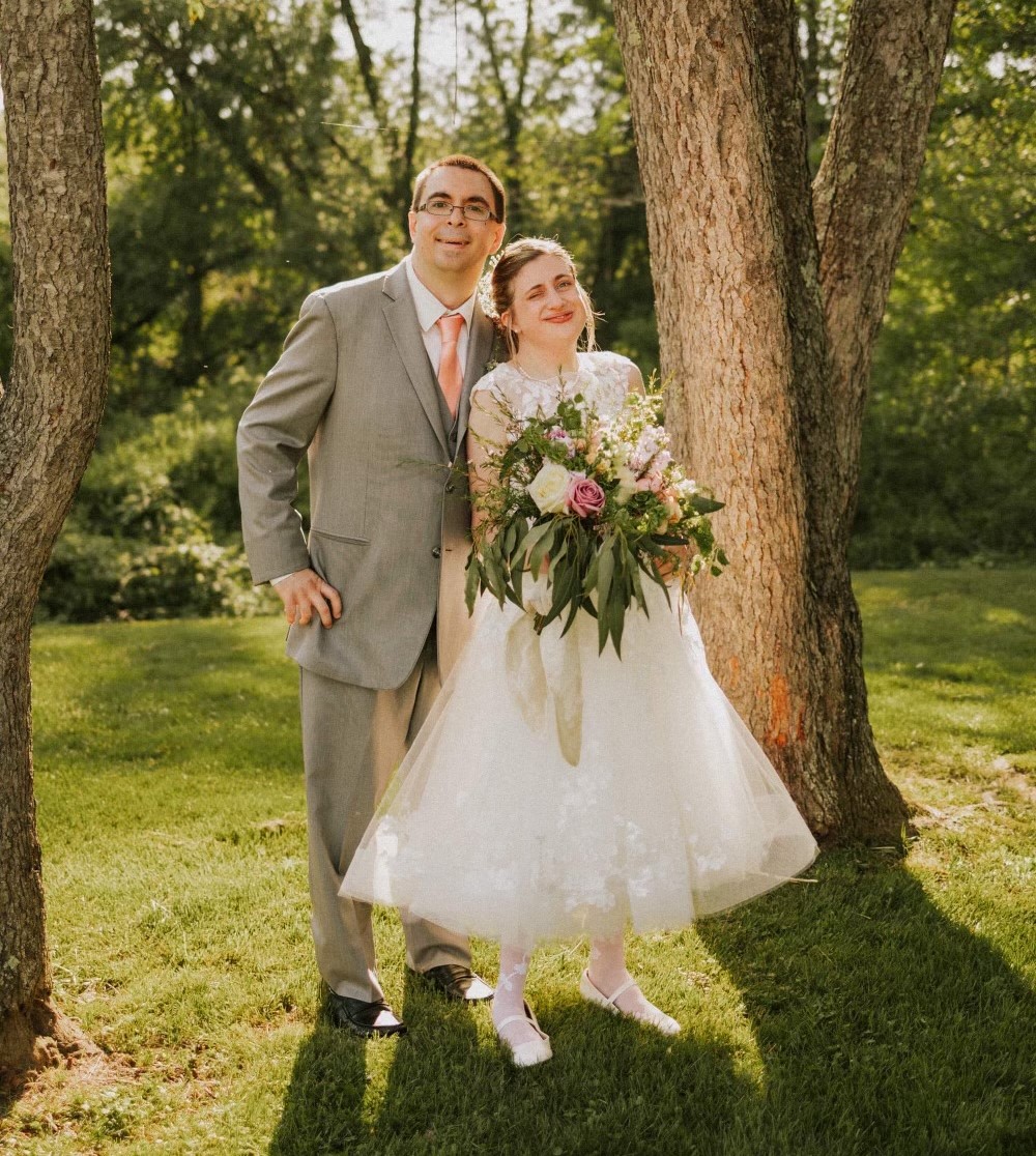 A couple on their wedding day. They are posed in front of a grassy area with trees around them and are wearing a wedding dress and suit. The bride is holding a bouquet of flowers.