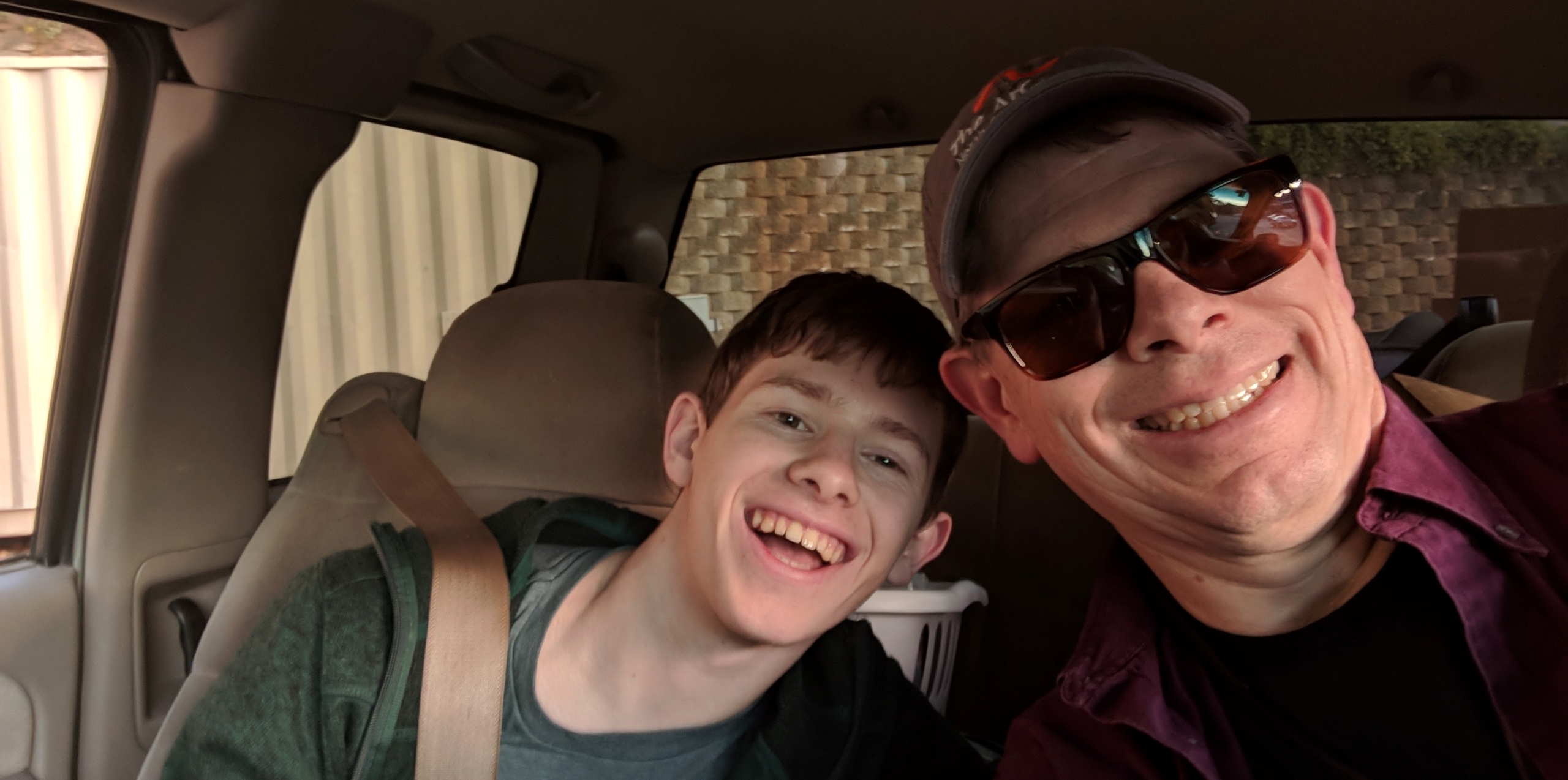 A selfie of a father and his teenage son, sitting in a car and smiling. The father is wearing sunglasses.