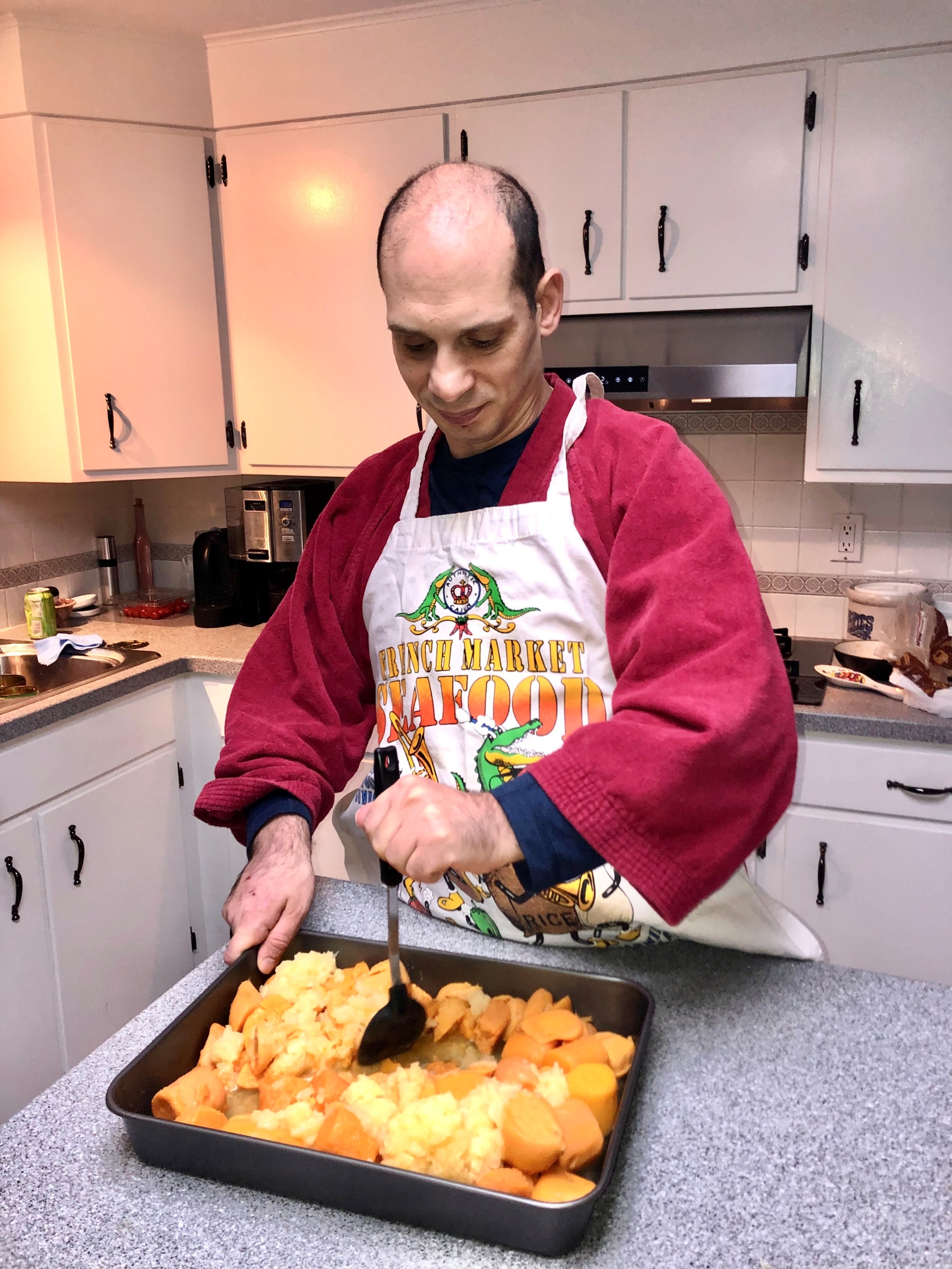 A middle-aged man stands in a kitchen with white cabinets. He is wearing a red shirt, and apron, and is using a cooking utensil on a sheet pan of food. 