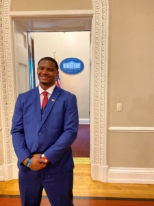 A man in a blue suit with a red tie stands in front of a doorway at the White House. In the background is a blue oval plaque on the wall with white text that reads "The White House" and behind him is an American Flag.