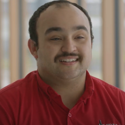 A man with a red shirt and mustache sits for an interview and smiles between sentences. He has dark brown hair and is balding slightly in the front of his head.