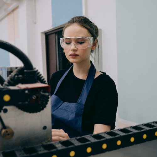 A white woman with dark hair in a bun stands in a factory setting. She has safety glasses on and is wearing a black t shirt and blue apron as she completes a task with some machinery in the foreground.