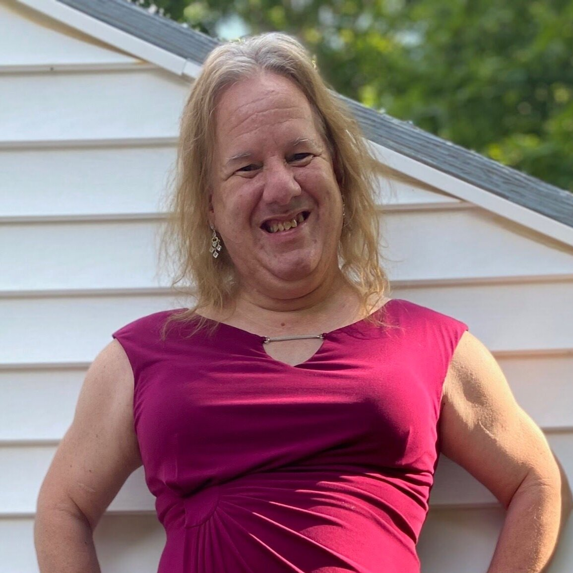 A woman in a bright pink sleeveless blouse stands smiling with her arms on her hips. She has blonde hair and behind her is the siding of a house. 