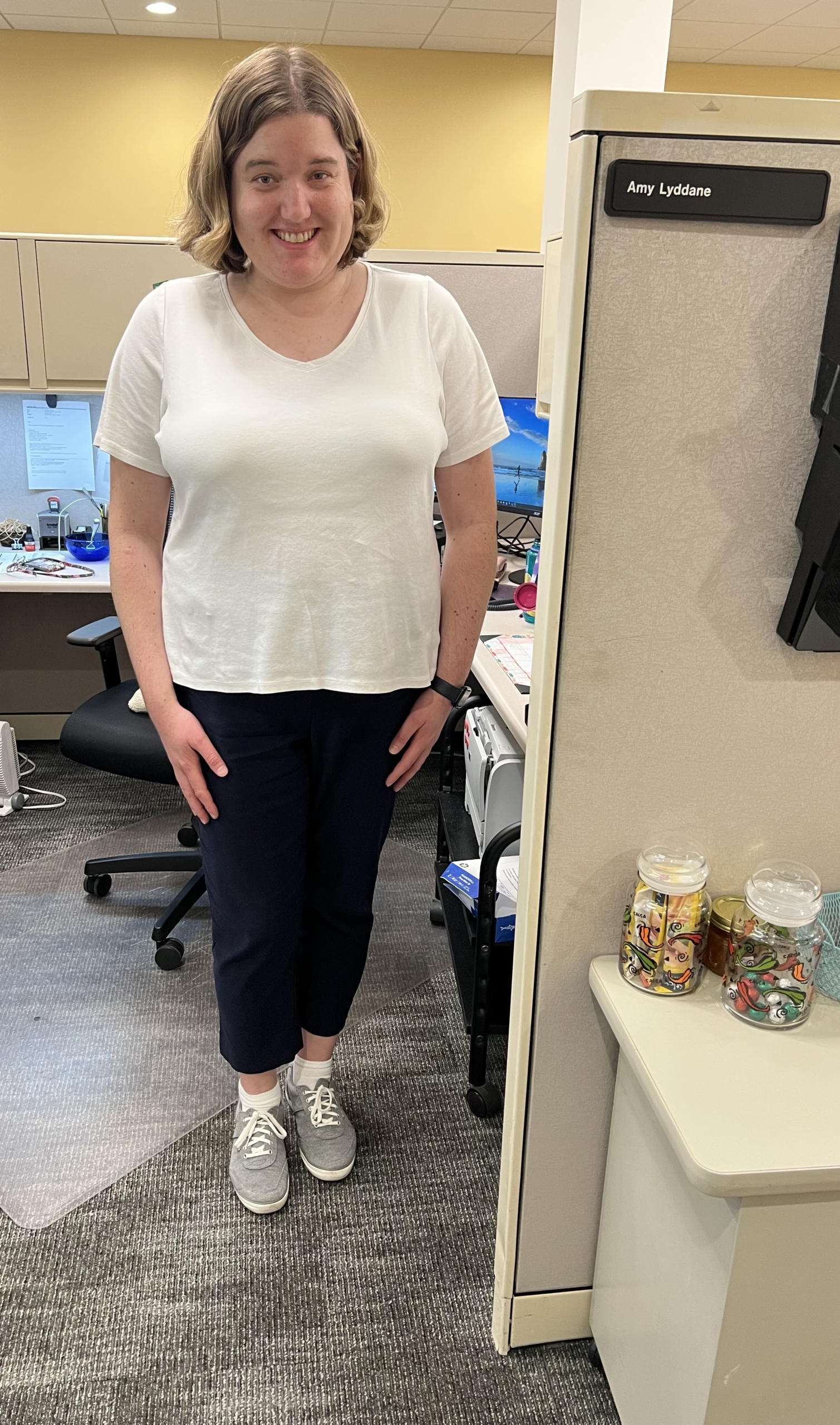 A white woman with blonde hair stands smiling with her arms at her sides in front of an office desk cubicle. She is wearing a white t shirt, black pants, and white shoes. 