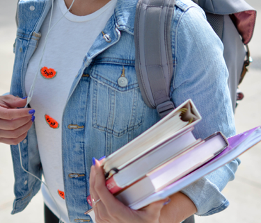 The torso of a young student wearing a light blue jean jacket over a white shirt. The student is holding three books and a notebook in their left hand and is wearing a backpack. You cannot see their face.