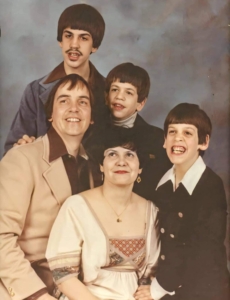 A professional photo of Roy and Arleen's family from 1976. Arleen is sitting in the middle and is wearing a tan dress. She has short, dark brown hair, red lipstick, and is wearing a gold necklace and earrings. Her husband, Roy, is on the left; he has shaggy brown hair and is wearing a light tan suit with a dark brown shirt underneath. Their three sons are seated behind them and to the right; they all have shaggy, bowl haircuts and dark brown hair. The young man behind Roy has a small moustache and is wearing a blue jacket. The young boy next to Roy is smiling and has a white turtleneck on. The young boy next to Arleen has a very big smile. He's wearing a blue jacket with a white collared shirt.