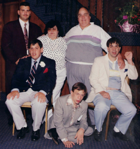 A photo from 1988 depicting a family of six. Three people are standing in the back, including a young adult man in a dark suit and purple tie; a woman wearing sunglasses, a white blouse with black polka dots, and white pants; and a man in a track suit. Two young men in suits are sitting in chairs in front of them. Another young man, also wearing a suit, is sitting cross-legged on the floor between them.
