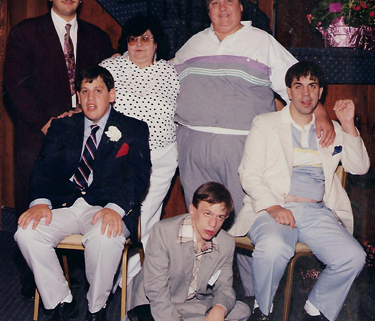 A photo from 1988 depicting a family of six. Three people are standing in the back, including a young adult man in a dark suit and purple tie; a woman wearing sunglasses, a white blouse with black polka dots, and white pants; and a man in a track suit. Two young men in suits are sitting in chairs in front of them. Another young man, also wearing a suit, is sitting cross-legged on the floor between them.