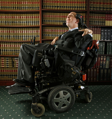 A man wearing a dark suit sits in a wheelchair. Behind him is a wall of books.