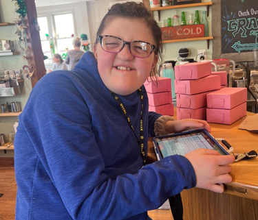 A young adult woman with developmental disabilities is holding a tablet. She's looking at the camera and has a big smile.
