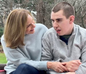 A young man with disabilities and his mom sit outside in a park. The mom is looking over at the son, holding his arm lovingly.