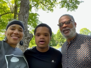 A Black family—mom, son, and dad—is standing outdoors. They are posing next to each other and smiling.