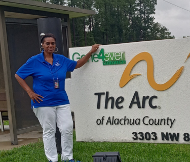 Betty Davis wears a blue top and white pants and stands outsie in front of The Arc of Alachua County sign.