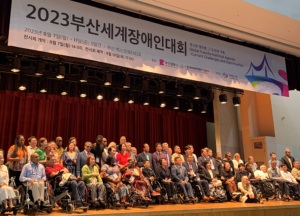 A large group of people with and without disabilities stand on stage at a conference.