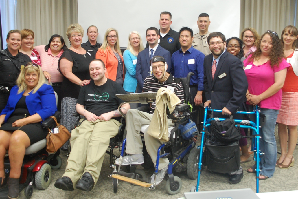 A group of people dressed in business attire poses for a photo. In the front row, there are three people in wheelchairs and one holding onto a walker.
